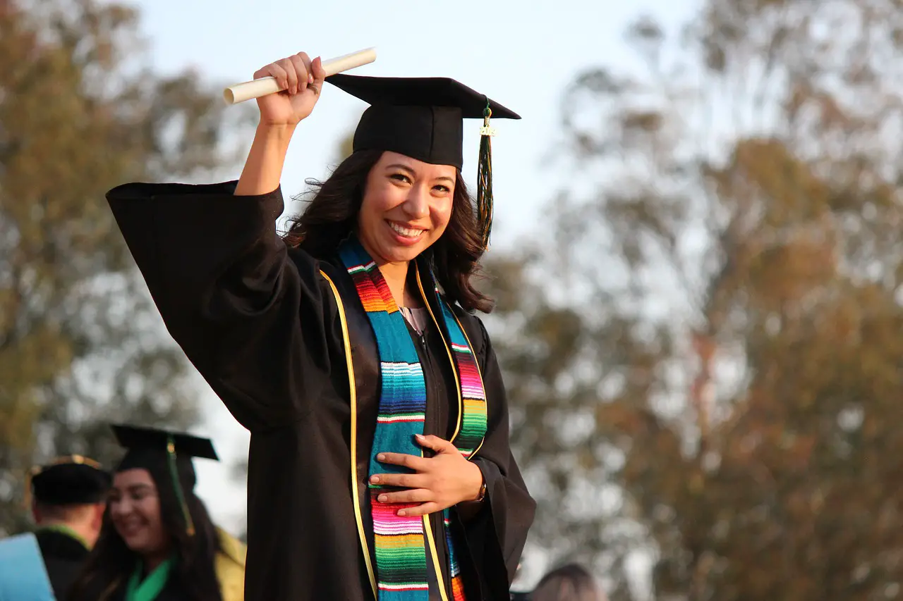 A female student in graduation regalia holding up her paper degree.
